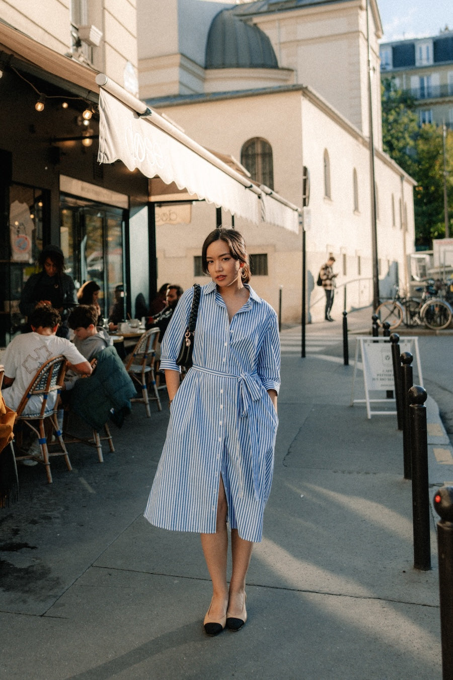 A woman wearing a blue and white vertically striped button-down shirt dress walks on a city sidewalk next to a cafe.